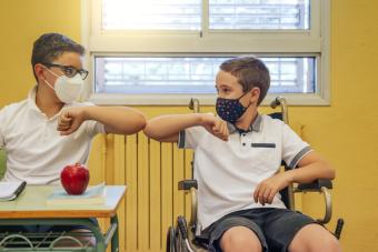 [image of two students in a classroom, one is seated at a desk and the other is in a wheelchair. They are giving each other an "elbow bump."]