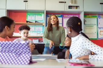 [image of a black female teacher leaning back on a desk in a classroom, surrounded by 4 seated adolescent students facing towards each other]